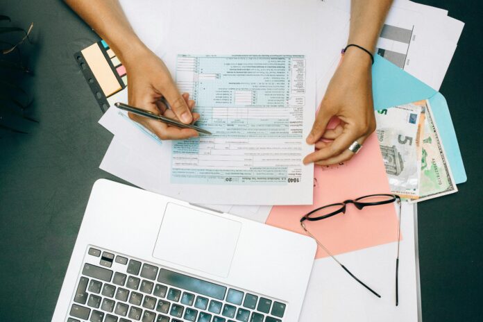 Pessoa preenchendo documento financeiro ao lado de notebook, dinheiro em espécie e óculos, representando organização financeira no dia a dia.