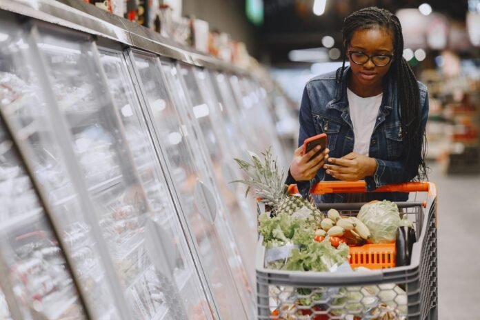 Mulher no corredor de um supermercado, com carrinho de compras cheio, consultando o celular para comparar preços dos produtos