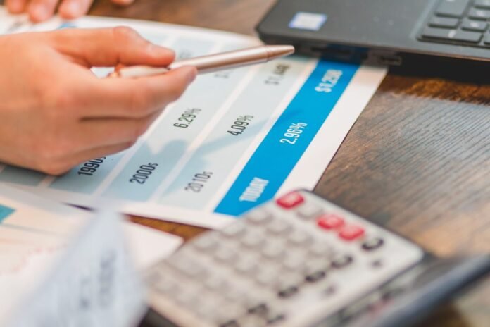 Mão segurando caneta aponta para tabela de taxas de juros ao lado de calculadora sobre mesa