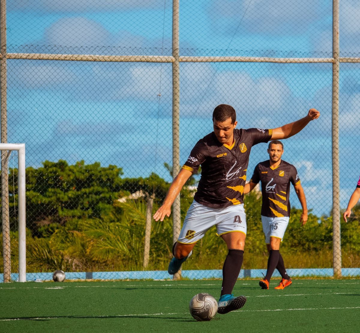 Jogador de futebol brasileiro chutando a bola em partida, camisa preta e dourada com área de patrocínio no peito