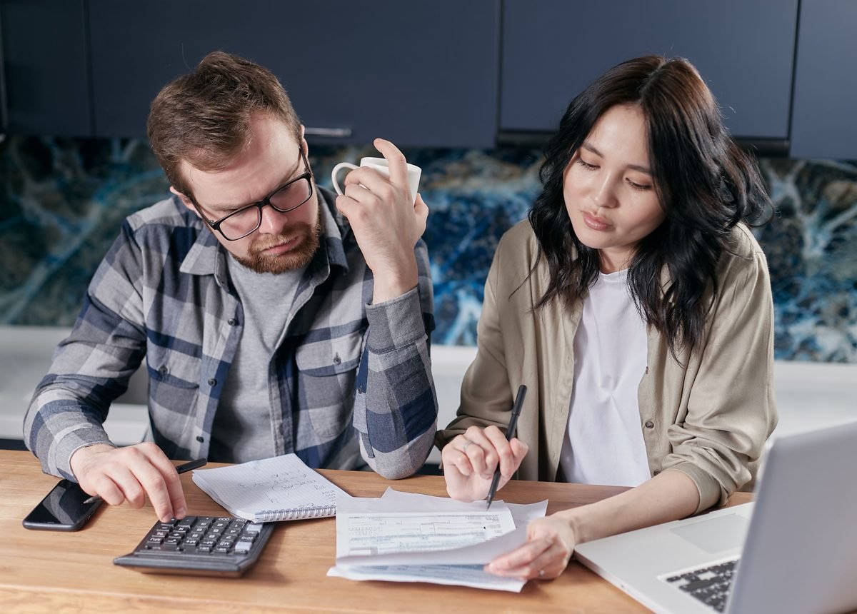 Casal organizando o orçamento doméstico em uma mesa de cozinha, com calculadora, caderno de anotações e laptop