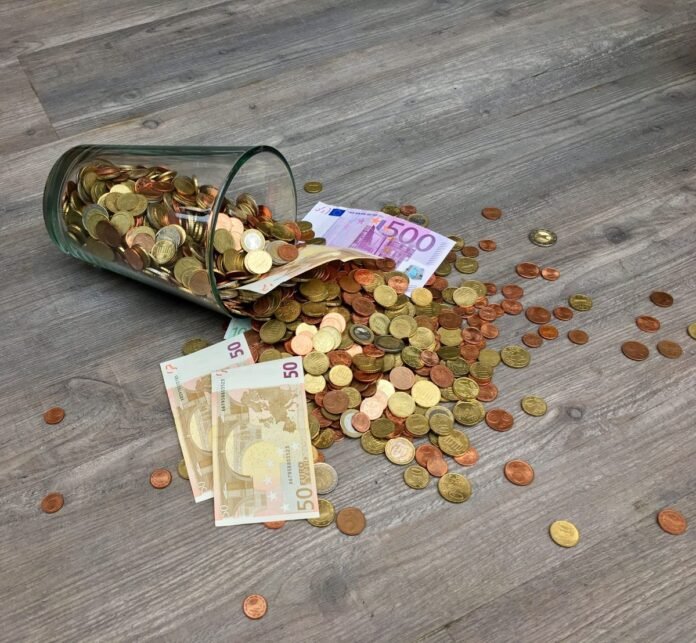 Pote de vidro tombado com moedas e cédulas de euro espalhadas sobre uma mesa de madeira, representando reserva de emergência