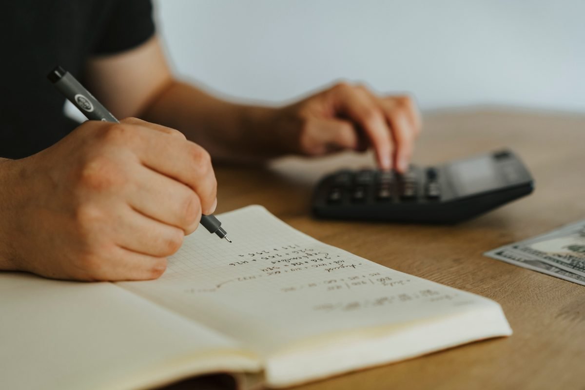 Mãos anotando cálculos em um caderno quadriculado ao lado de uma calculadora, em um planejamento financeiro
