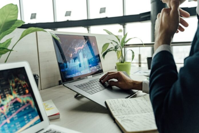 Profissional analisando gráficos de investimentos em um notebook, em mesa de escritório organizada, com caderno de anotações e plantas ao fundo
