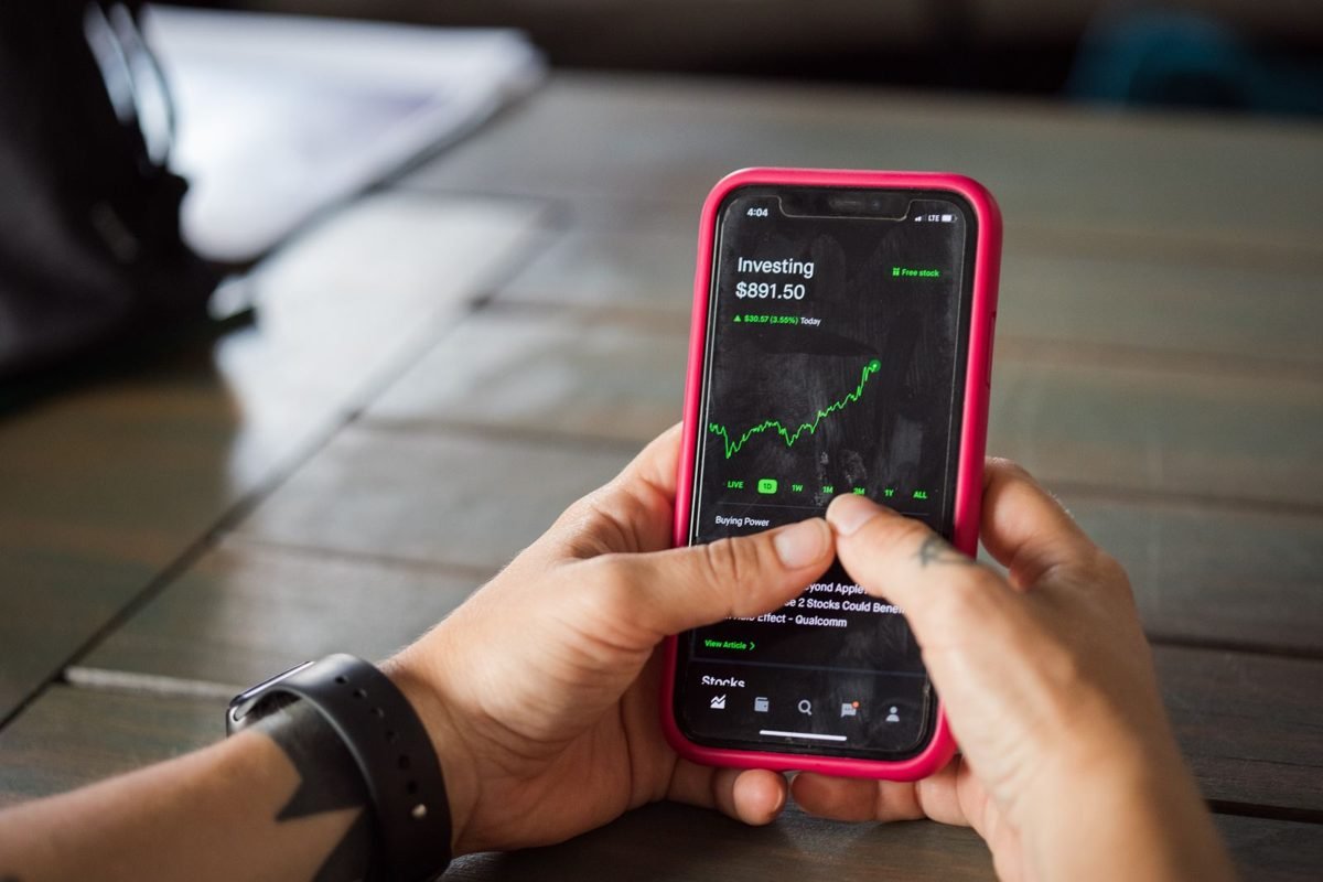 Mãos segurando um smartphone com capa rosa que exibe um aplicativo de investimentos com gráfico verde em alta
