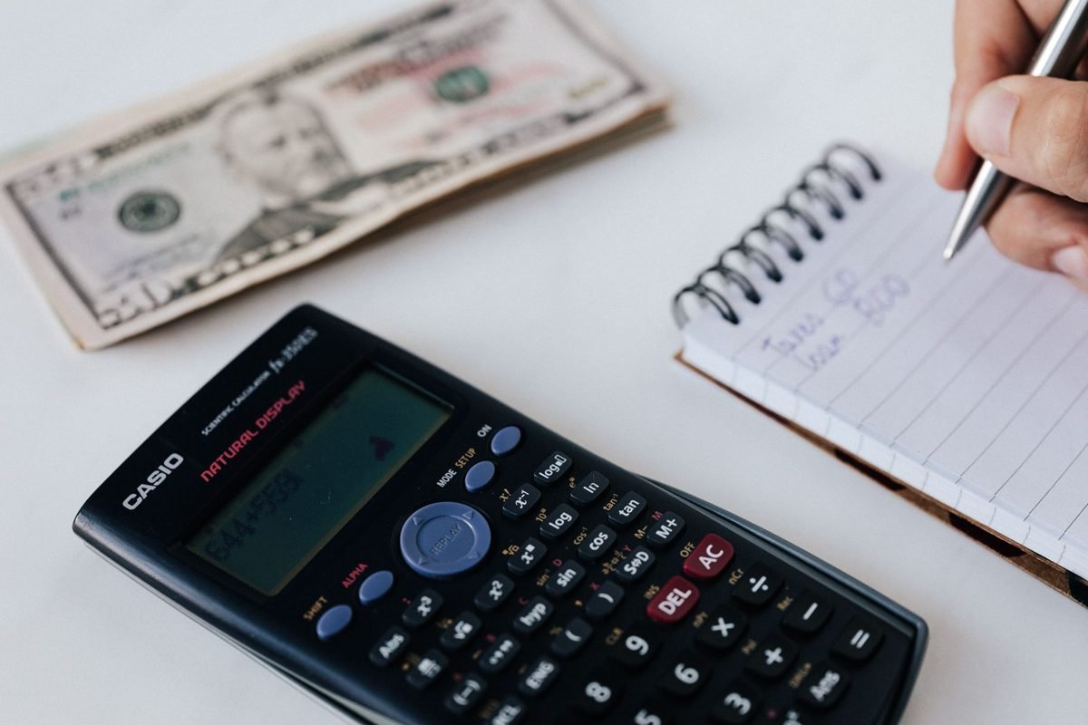 Calculadora científica sobre a mesa, ao lado de notas de dólar e de uma mão anotando valores em um caderno espiral