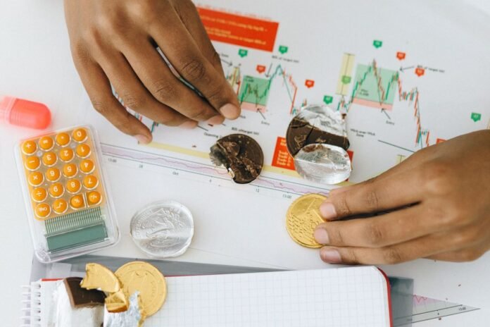 Mãos sobre um gráfico impresso de variação de preços, com moedas, uma calculadora laranja e um caderno de anotações sobre a mesa