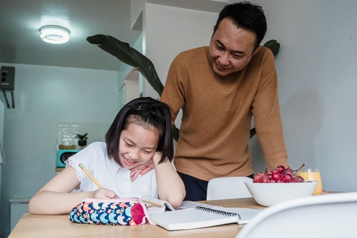 Pai sorrindo em pé ao lado da filha, que escreve em um caderno sobre a mesa da cozinha