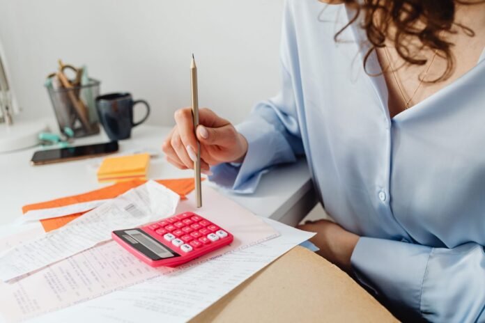Mulher com um lápis na mão calculando despesas em uma calculadora rosa, sobre uma mesa com boletos e comprovantes