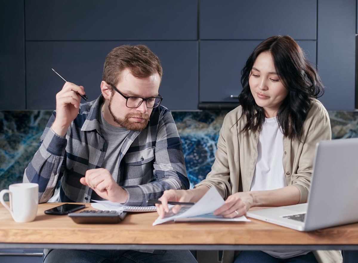 Casal sentado à mesa de madeira analisando papéis, com calculadora, celular, caderno, xícara e notebook sobre a mesa