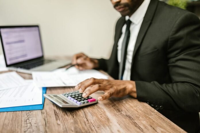 Homem de terno sentado à mesa usando uma calculadora, com documentos financeiros e um notebook à frente