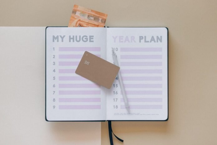Cartão de crédito dourado sobre uma agenda aberta com lista numerada, ao lado de uma caneta e de uma nota de dinheiro