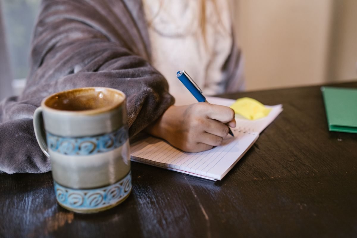 Mão escrevendo em um caderno de folhas pautadas ao lado de uma caneca de café sobre uma mesa escura